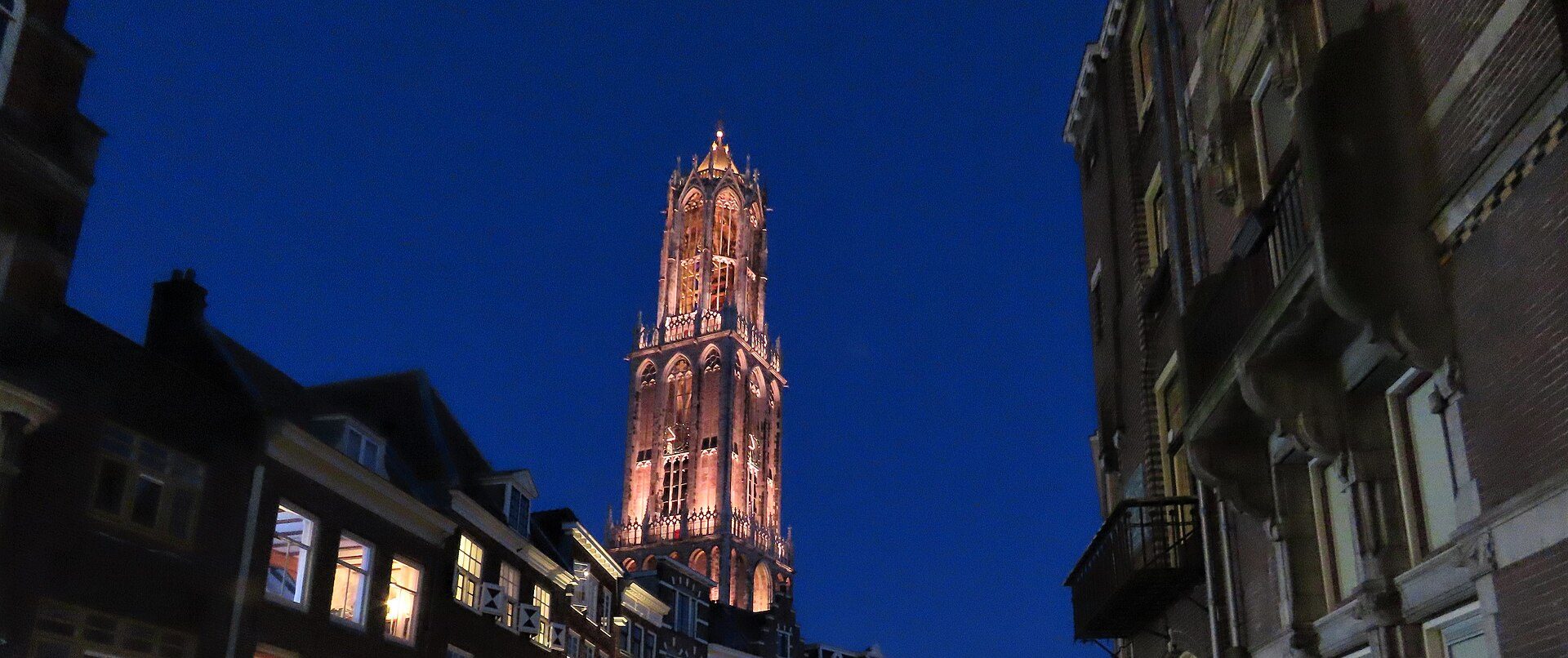 De verlichte Domtoren in Utrecht in de avondschemering, tussen oude grachtenpanden